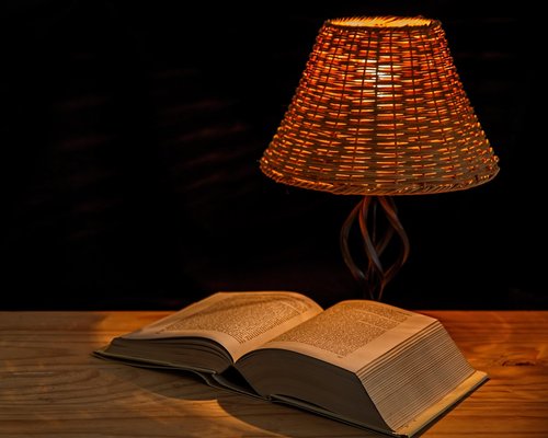 A person reading a printed book at night under a warm, soft reading lamp
